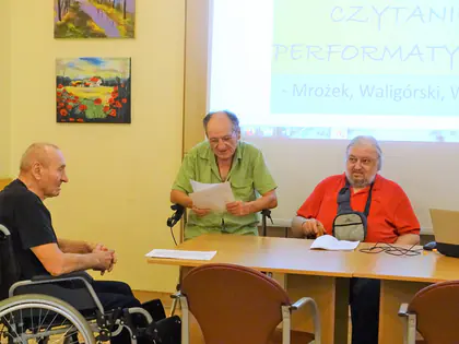 Mieszkańcy czytają teksty przy stole, na ekranie napis Czytanie performatywne