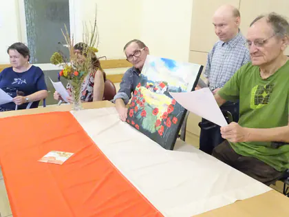 Mieszkańcy czytają wiersze przy stole z biało-czerwonym obrusem i obrazem z makami