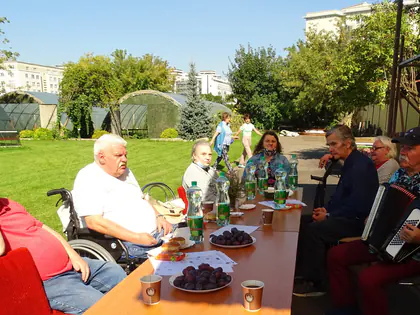 Mieszkańcy siedzą przy stole w ogrodzie, jeden gra na akordeonie