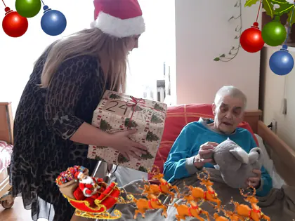 Kobieta w czapce Mikołaja wręcza paczkę mieszkance leżącej w łóżku