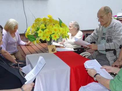 Seniorzy siedzą przy stole, który ozdobiony jest flagą Polski i żółtym bukietem kwiatów