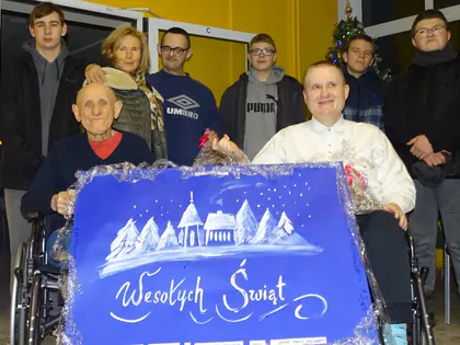 Młodzież i mieszkańcy trzymają ręcznie malowany plakat z napisem Wesołych Świąt