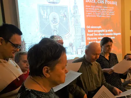 Mieszkańcy śpiewają pieśni patriotyczne z kartek przed ekranem z tekstem pieśni Boże, coś Polskę
