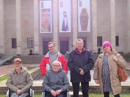 Mieszkańcy, dwóch na wózkach, i opiekunka pozują przed gmachem Muzeum Narodowego w Warszawie