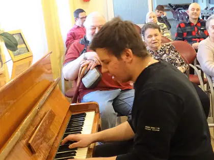 Młody mężczyzna gra na pianinie, mieszkańcy słuchają