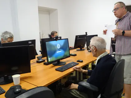 Prowadzący zajęcia komputerowe stoi przy seniorach siedzących przy monitorach