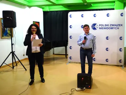 Kobieta czyta zapowiedź z kartki, obok mężczyzna śpiewa do mikrofonu na tle ścianki Polskiego Związku Niewidomych