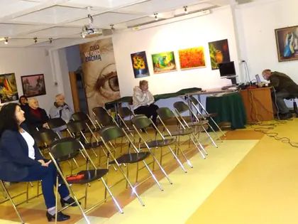 Sala z obrazami na ścianach i publicznością na krzesłach podczas koncertu
