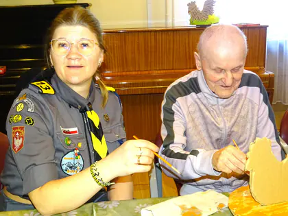 Harcerka w mundurze i starszy mężczyzna malują kartonową kurę, w tle pianino