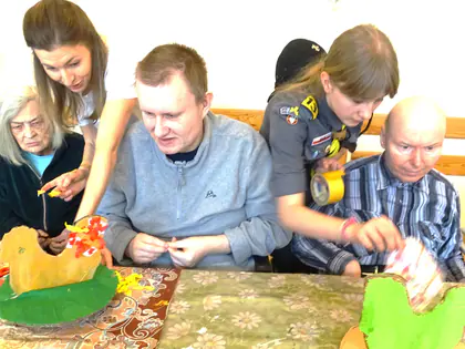 Mieszkańcy i harcerki ozdabiają przy stole kartonowe kury, jedna z nich trzyma taśmę