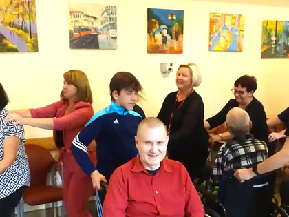 Uczestnicy tańczą w kole, na pierwszym planie siedzi mężczyzna w czerwonej koszuli