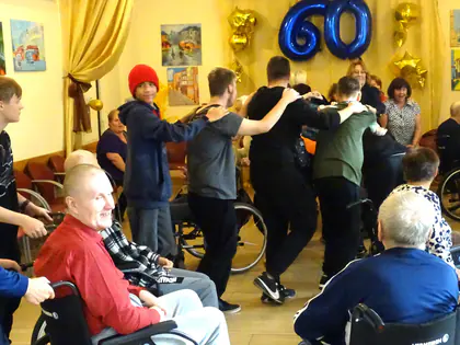 Uczestnicy zabawy tańczą pociągiem w sali z balonem w kształcie liczby 60, mieszkańcy siedzą na wózkach