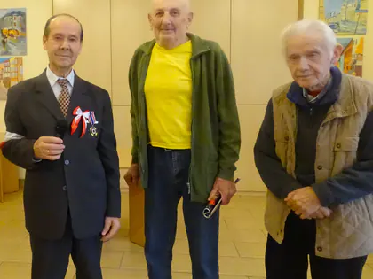 Trzech starszych mężczyzn stoi w sali, jeden w garniturze z mikrofonem i biało-czerwoną kokardą