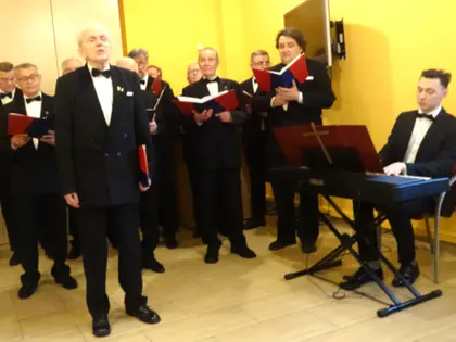 Chór męski w frakach śpiewa z nutami w czerwonych teczkach, obok pianista przy keyboardzie