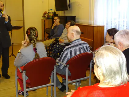 Mężczyzna w garniturze śpiewa do mikrofonu przed siedzącymi mieszkańcami
