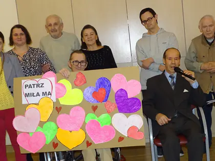 Mieszkańcy i goście pozują do zdjęcia z kolorowymi papierowymi sercami, pośrodku mężczyzna z mikrofonem