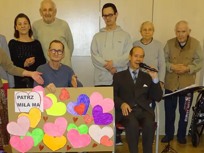 Grupa mieszkańców i gości stoi za tablicą z napisem Patrz miła ma i kolorowymi sercami, śpiewak z mikrofonem siedzi pośrodku