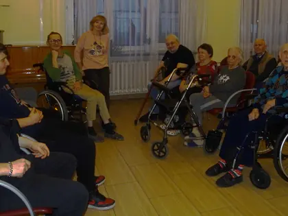 Młodzi goście i mieszkańcy siedzą w sali, część na wózkach i z balkonikami