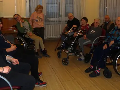 Młodzież i mieszkańcy siedzą w sali na krzesłach i wózkach
