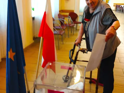 Starsza kobieta z balkonikiem wrzuca kartę do przezroczystej urny, obok flagi Polski i Unii Europejskiej