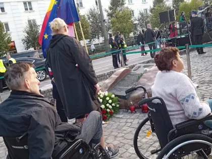Dwie osoby na wózkach przy pomniku z kwiatami podczas uroczystości