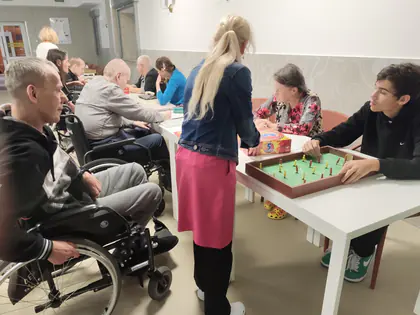 Przy stole mieszkanka i chłopiec grają w piłkarzyki, obok stoi kobieta i siedzą inni uczestnicy