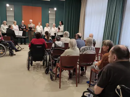 Zespół wokalny w jasnych strojach śpiewa przed siedzącymi mieszkańcami, część na wózkach