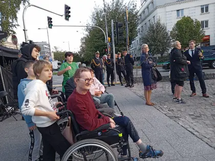 Chłopiec i mężczyzna na wózku wśród uczestników uroczystości na ulicy, w tle poczet sztandarowy