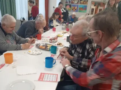 Mieszkańcy i goście grają w bingo przy długim stole z kawą i ciastem