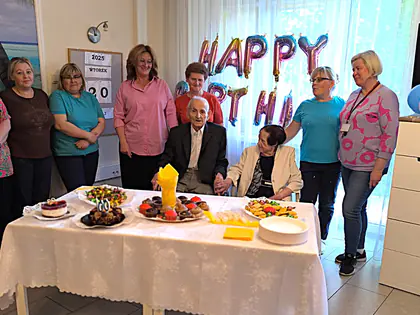 Pracownice stoją za stołem z tortem i owocami obok jubilata i jego żony, na ścianie balony z napisem Happy Birthday