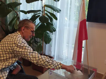 Mężczyzna wrzuca kartę do przezroczystej urny wyborczej, obok stoi biało-czerwona flaga