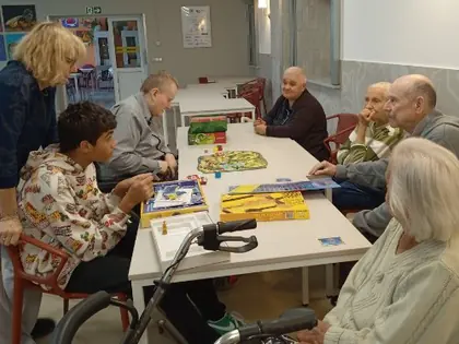Grupa osób przy stole z grami planszowymi, jeden z chłopców pochyla się nad planszą