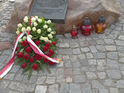 Głaz z tablicą pamiątkową, przy nim biało-czerwone kwiaty i znicze