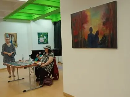 Autorka siedzi przy stoliku z mikrofonem w galerii, obok stoi kobieta, na ścianie kolorowy obraz