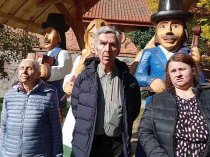 Mieszkańcy i opiekunka stoją pod drewnianą wiatą z rzeźbionymi postaciami w skansenie