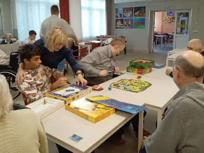 Mieszkańcy i młodzież przy stołach grają w gry planszowe, obok stoi kobieta