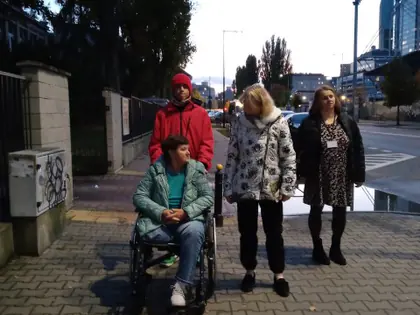 Pracownicy i mieszkanka na wózku idą chodnikiem, w tle osiedlowe budynki