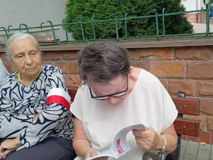 Dwie starsze kobiety siedzą na ławce, jedna ma biało-czerwoną opaskę, druga czyta broszurę
