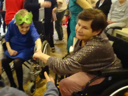Uczestnicy balu w maskach tańczą na sali, część siedzi na wózkach