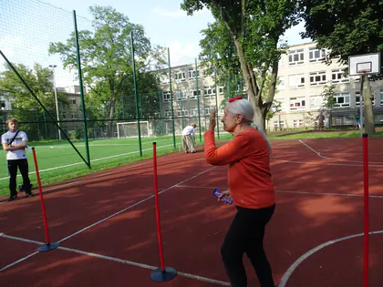 Starsza kobieta w pomarańczowej bluzie przechodzi tor przeszkód na boisku