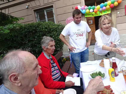 Mieszkańcy przy stole z poczęstunkiem przed budynkiem z balonami