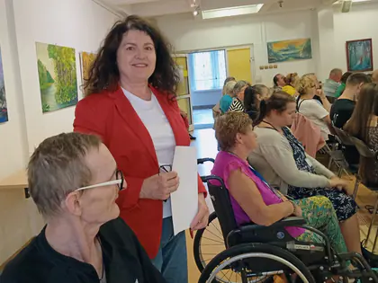 Prelegentka w czerwonej marynarce stoi przed publicznością w sali wystawowej z obrazami