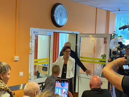 Korytarz z zegarem na ścianie, w drzwiach goście, na pierwszym planie uczestnicy i osoba z telefonem