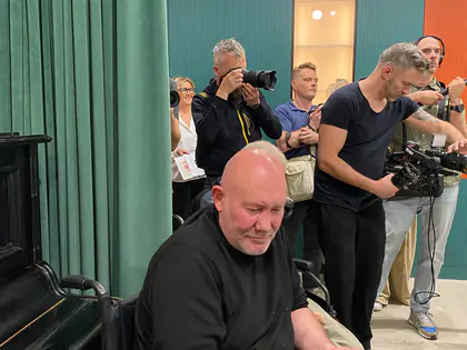 Mężczyzna na wózku klaszcze, za nim fotoreporterzy i operator kamery