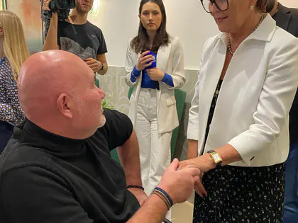 Kobieta w białym żakiecie ściska dłoń mężczyzny na wózku, w tle operator z kamerą