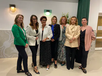 Siedem kobiet w eleganckich strojach stoi w rzędzie na tle jasnej ściany