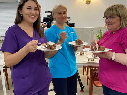 Trzy kobiety w kolorowych fartuchach jedzą ciasto z talerzyków