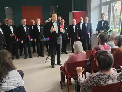 Dyrygent i chór męski w czarnych garniturach występują przed siedzącymi słuchaczami