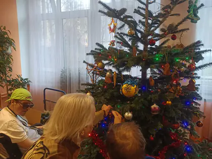 Dwie osoby przy choince zawieszają ozdoby, z boku siedzi mężczyzna w zielonej czapce