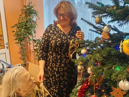 Kobieta zawiesza ozdobę na choince, obok siedzi starsza mieszkanka